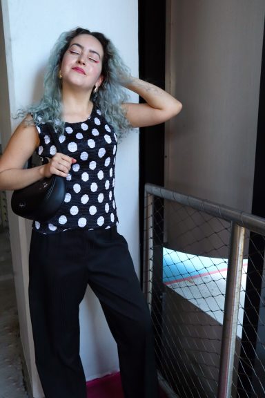 Young woman with blue hair in a polka dot top, holding a purse, standing by a railing.